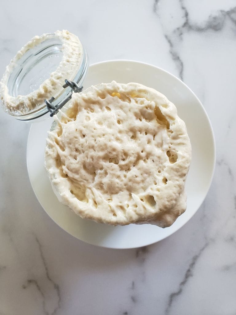 A birds eye view of of an active sourdough starter billowing out the top of an open flip top jar. It is sitting on a white ceramic plate and the starter is maintaining its height and isn't sinking back into the jar. It is pillowy and airy. 