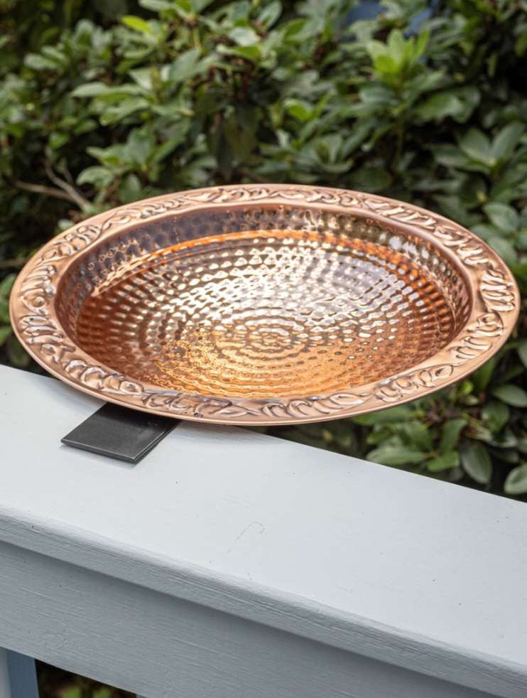 An outdoor deck railing with a copper birdbath attached to the top railing. Using bird baths will help attract birds to your garden. 