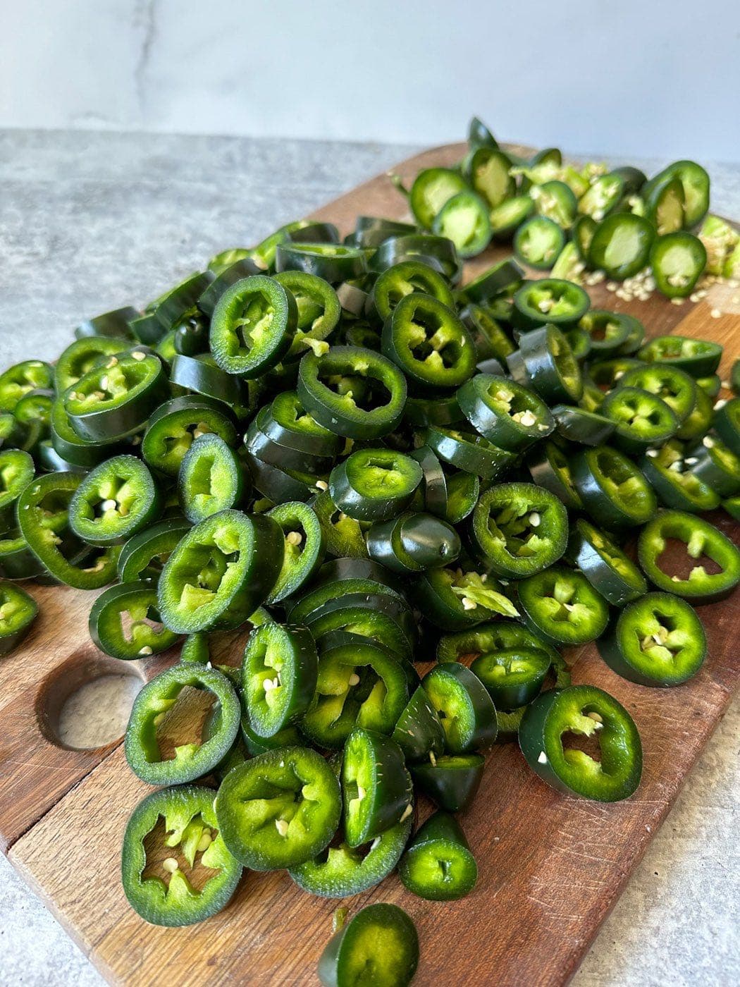 A mound of slices of jalapeños are on a cutting board, on the other side of the cutting board are the stem ends with some of the membrane and seeds left behind.   