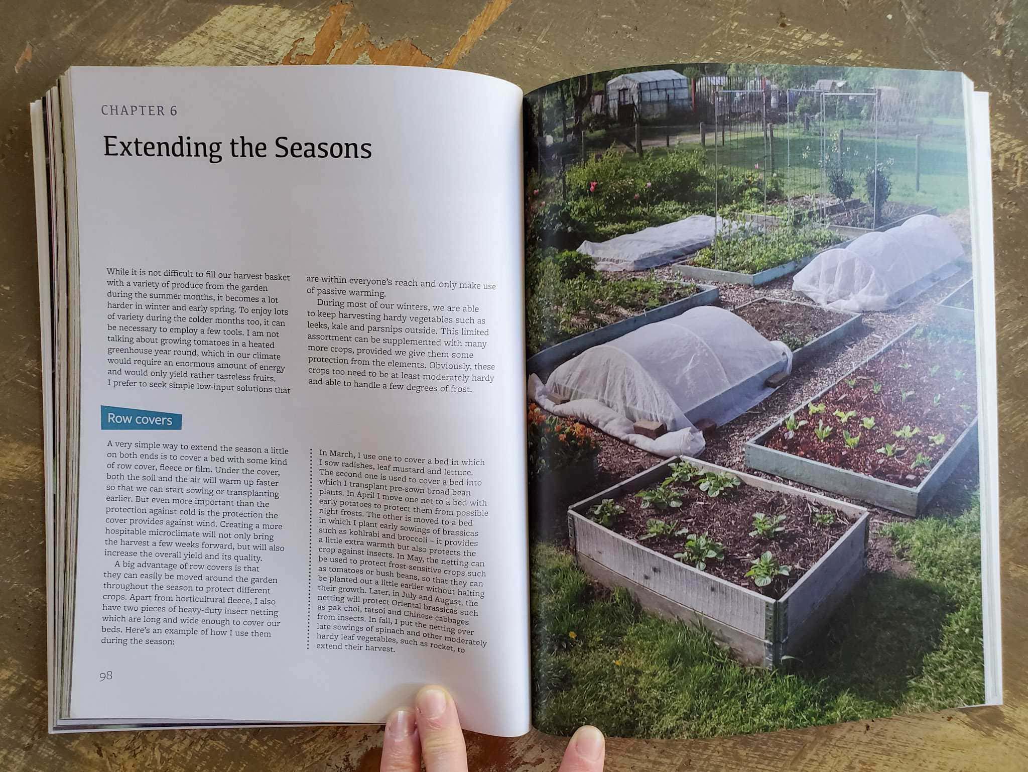 A gardening book is open to a section about "Extending the Seasons". One page is a full photograph of garden beds, some are left uncovered while others are covered in row covers, and some have wire trellises.  