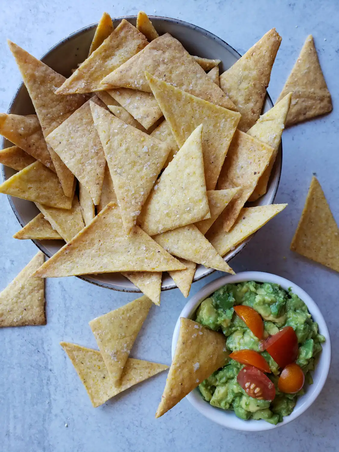 A close up image of a bowl of sourdough corn chips. They are golden to golden brown in color and triangular in shape. Specks of sea salt are visibly baked into the top of the chips. The bowl is overflowing with a few chips scattered around the perimeter of the bowl and a smaller white ramekin of fresh guacamole garnished with halved cherry tomatoes sits nearby. 