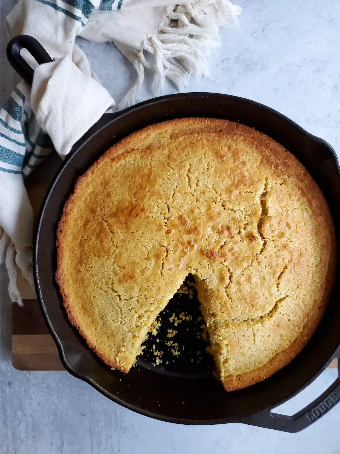 Sourdough cornbread after it has baked sitting in a cast iron skillet on top of a wood cutting board. A slice has been cut out of the cornbread as if it were a pie, leaving an empty triangular space behind, showing the black cast iron below.
