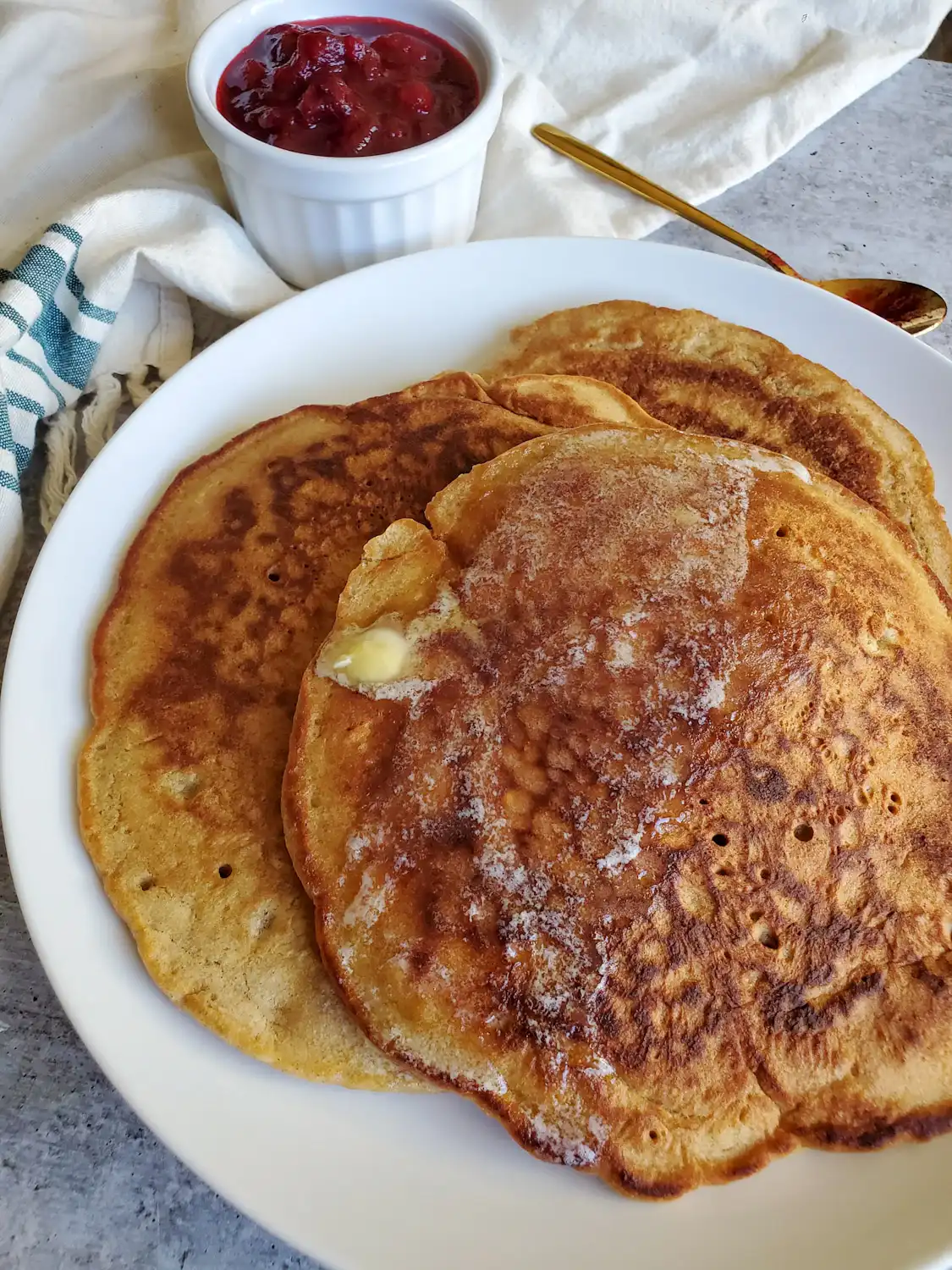 A stack of sourdough pancakes sit atop a white ceramic plate. A pad of butter is sitting on the top pancake and is shrinking in size as it melts throughout the top portion of pancake. A ramekin of fresh cranberry sauce sits in the background next to a gold spoon. Use this sourdough discard recipe to make delicious pancakes. 