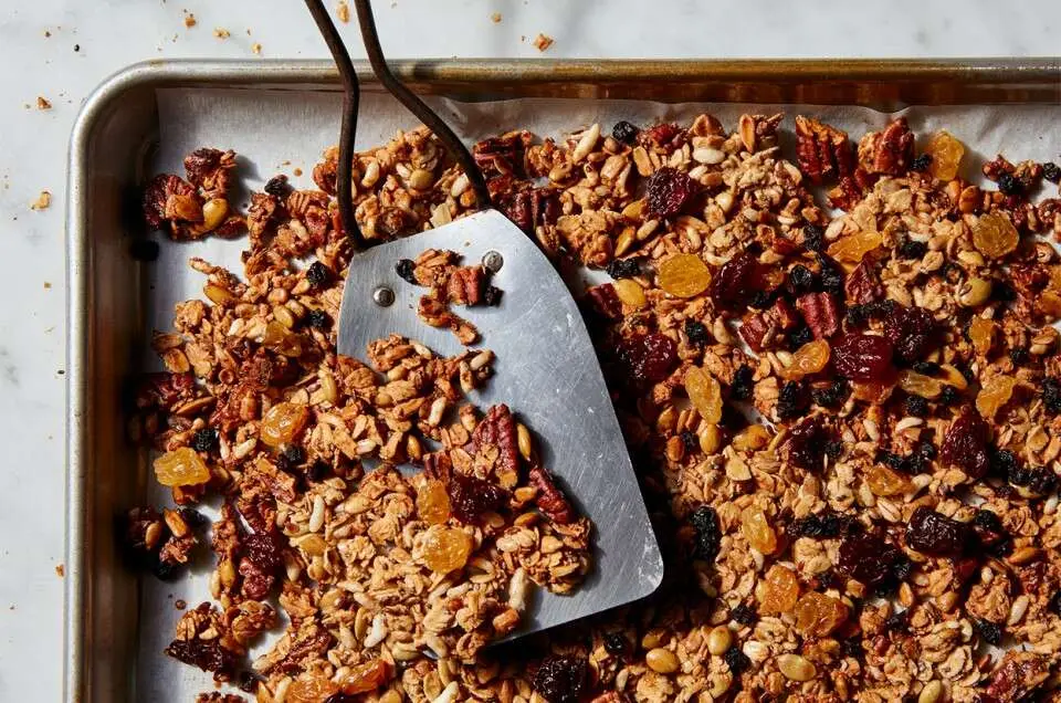 A baking sheet is full of homemade granola, a mixture of nuts, oats, and dried fruit are lightly brown in color. A flat spatula is sitting on the sheet pan, partially covered in granola. 