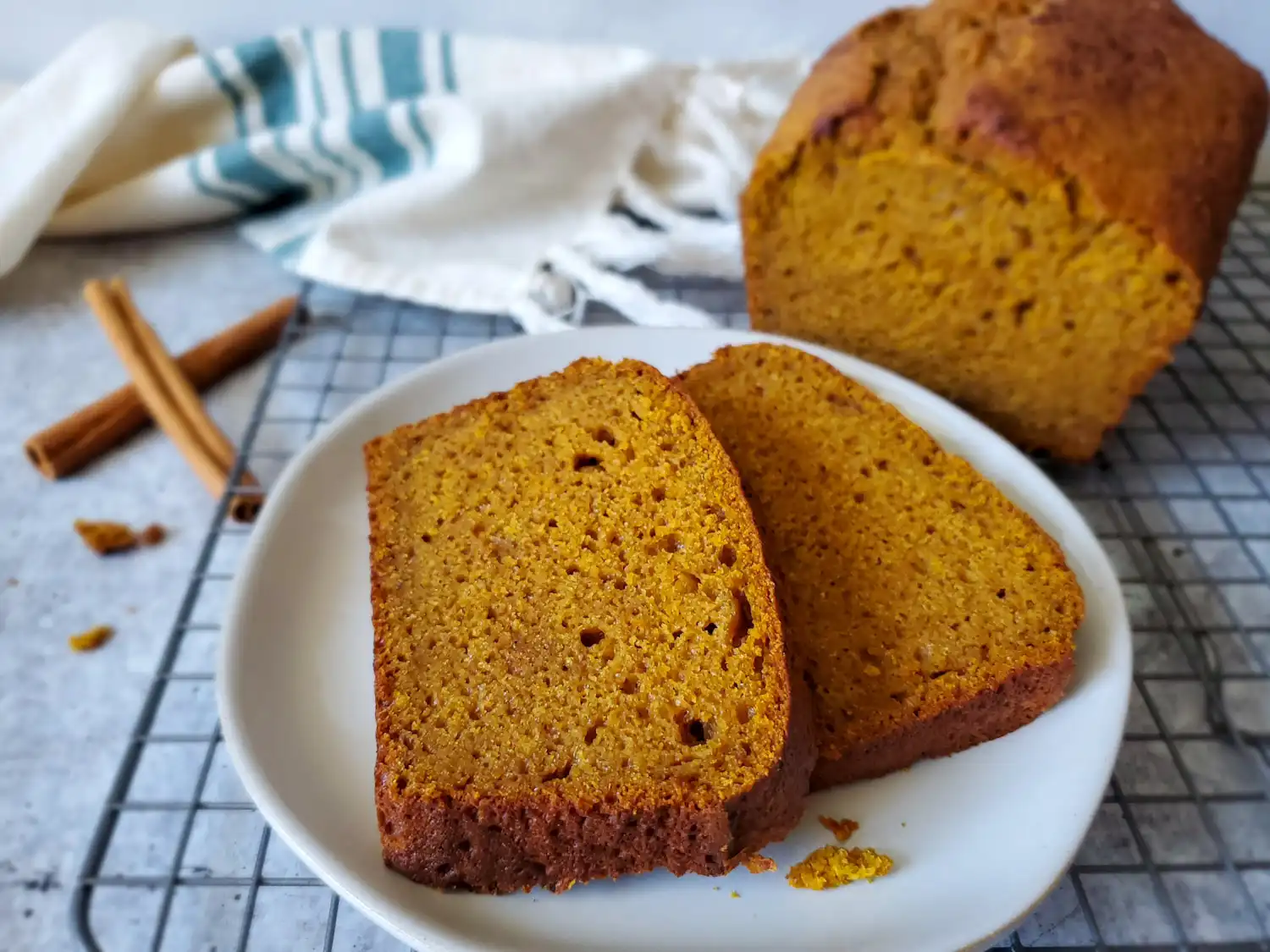 Two thick slices of sweet bread on a small plate. The bread is slightly orange and brown in color, sourdough pumpkin spice bread. The plate is resting on a cooling rack, and the cut loaf of pumpkin bread is blurry in the background with crumbs scattered around. 