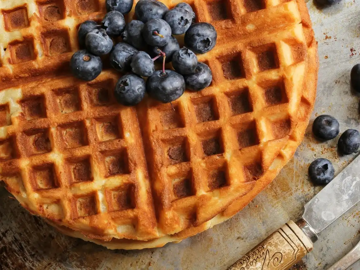 A close up image of a lightly brown waffle with blueberries on top of it. A few loose blueberries garnish the area around the waffle along with an antique butter knife.