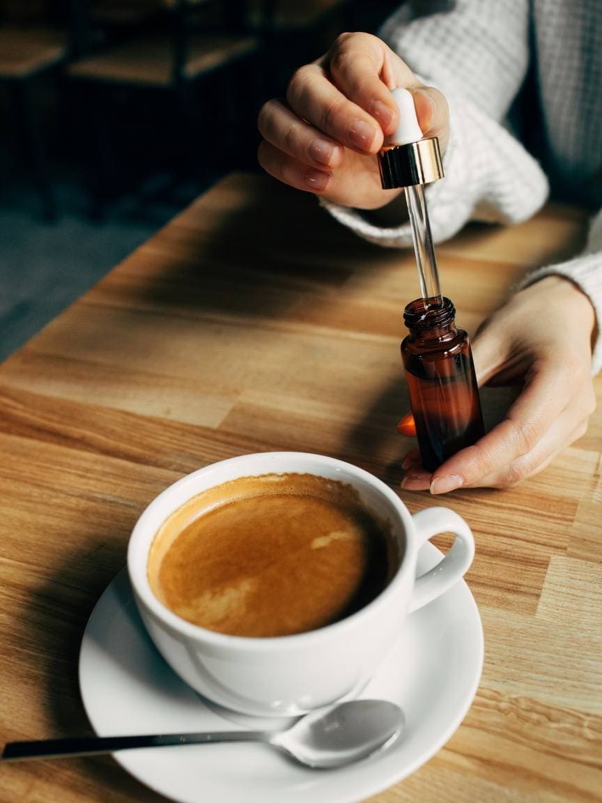 A coffee drink is shown in a white coffee cup sitting on a white plate. A person is inserting a dropper into an amber colored bottle to add oil to their coffee. When you choose CBD oil, you can take it in a variety of ways. 