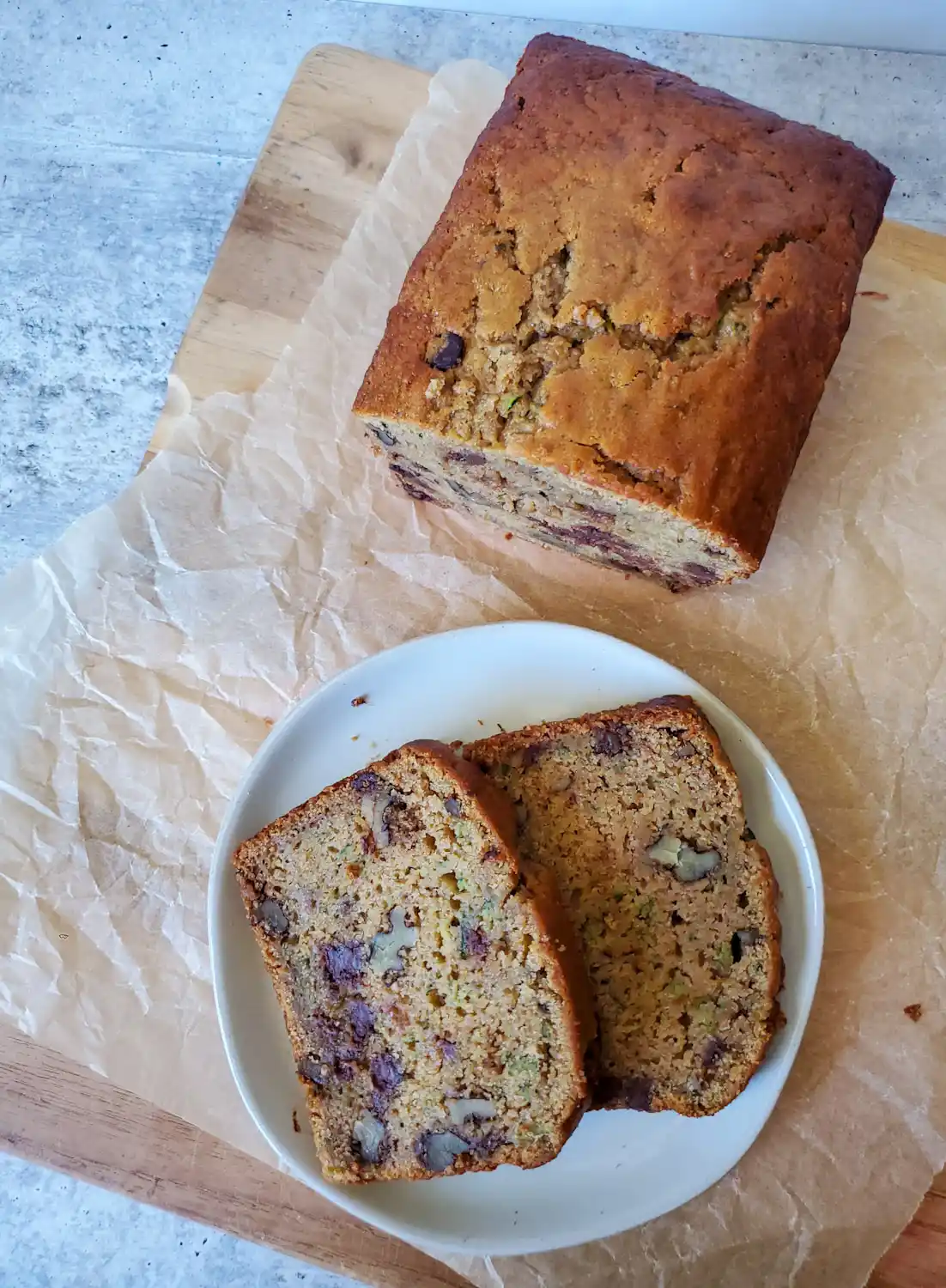A birds eye view of two slices of sourdough zucchini bread with walnut and chocolate chips that are sitting on a white ceramic plate. Underneath is a piece of parchment paper and the remaining loaf of bread that has yet to be cut. 
