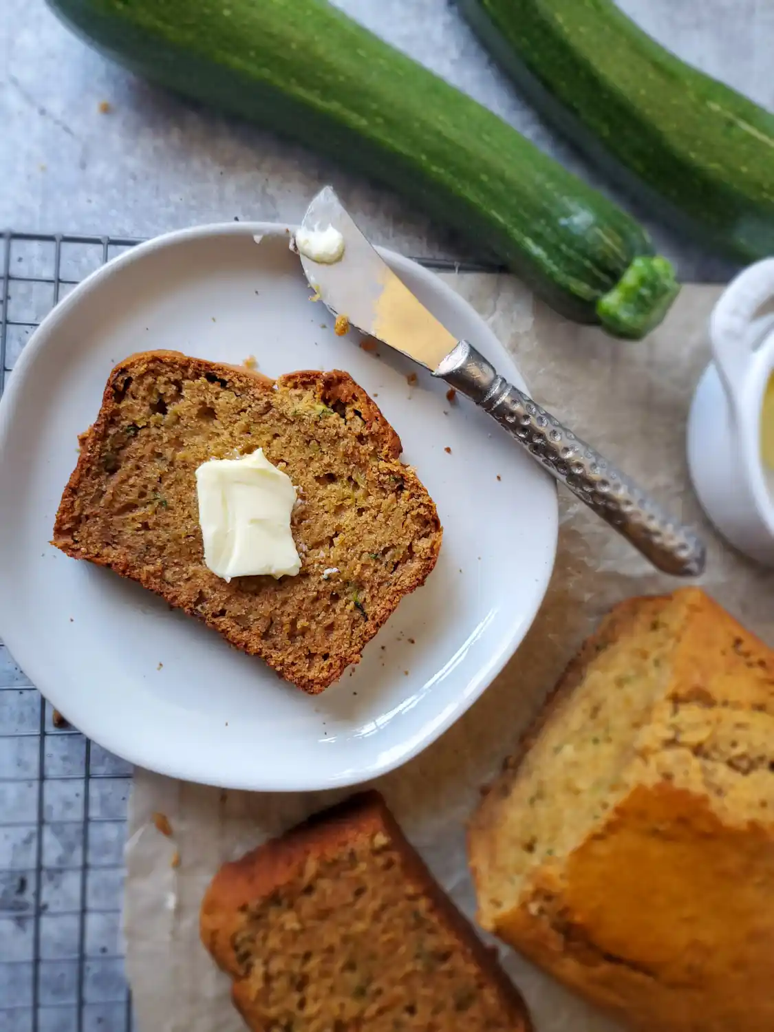 A slice of sourdough zucchini bread with a tab of butter sitting in the middle of the slice on a small ceramic plate,  a small butter knife is resting on the plate. Nearby is the rest of the loaf as well as some fresh summer squash. Using extra zucchini for sourdough discard recipes is a great way to use up a surplus harvest.