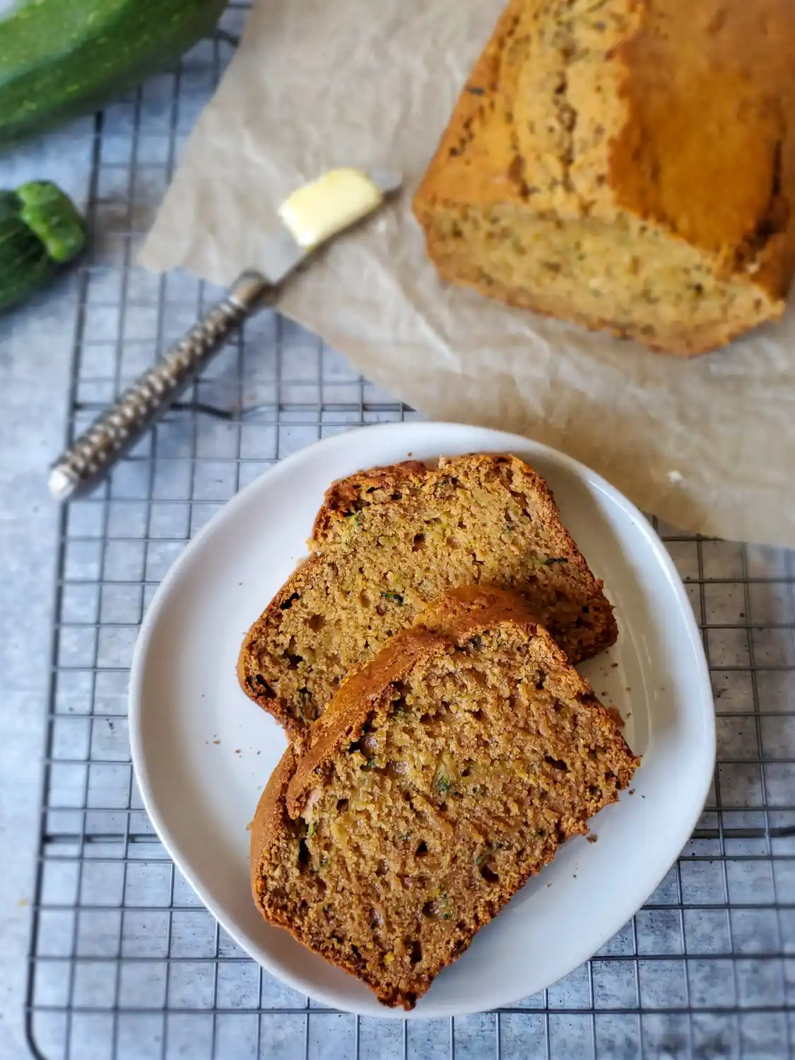 Two slices of sourdough zucchini bread sit atop a white ceramic plate. Nearby is the rest of the loaf sitting on parchment paper along with a small butter knife with a tab of butter resting on the end. Some squash is flanking the edge of the image for garnish. 