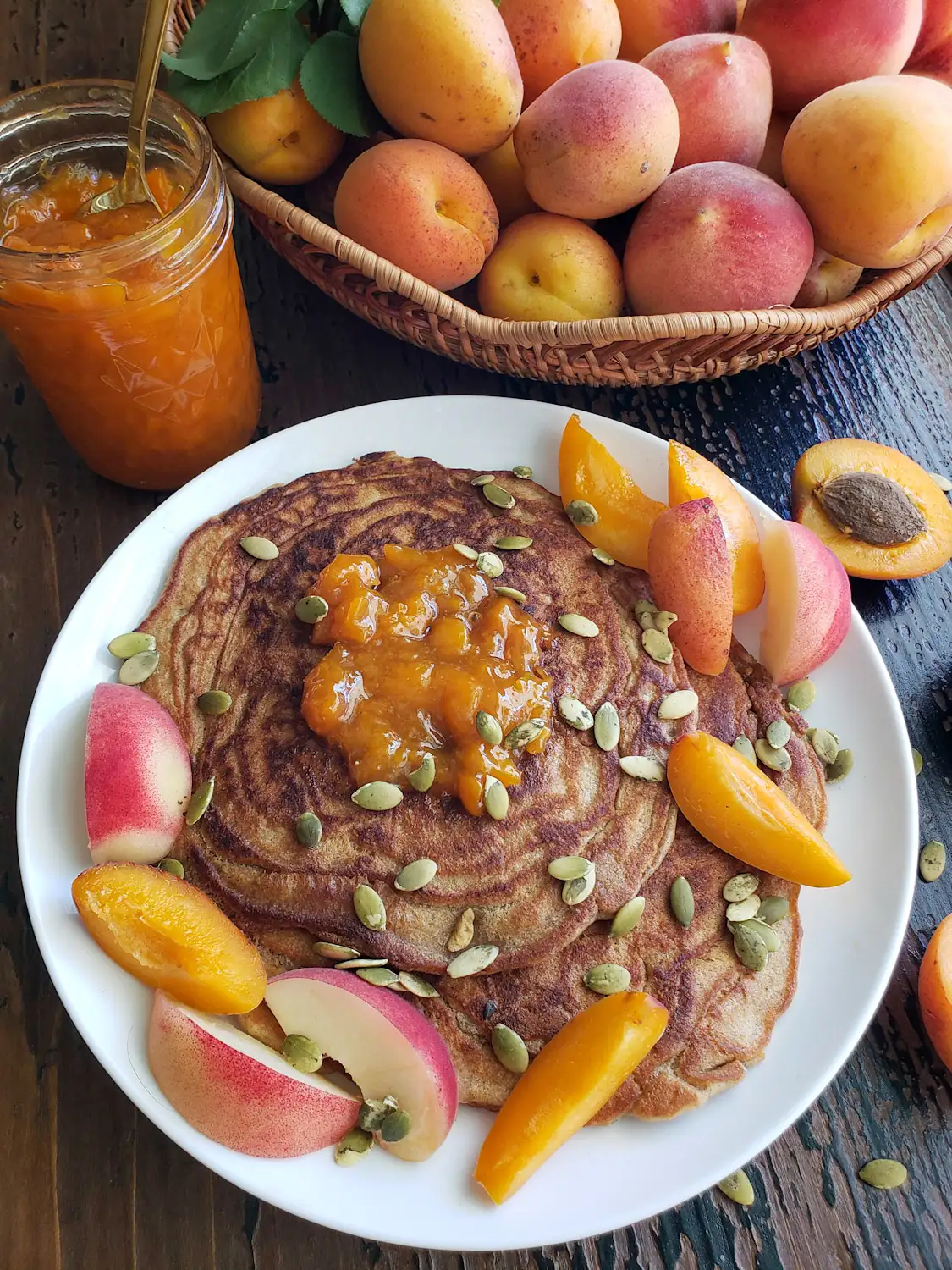 Two sourdough pancakes are stacked onto a white ceramic plate with a dollop of low sugar peach jam on top along with pumpkin seeds, slices of apricots, and slices of white peach. A half pint jar of jam is just above the plate along with a wicker basket of fresh apricots and peaches. 