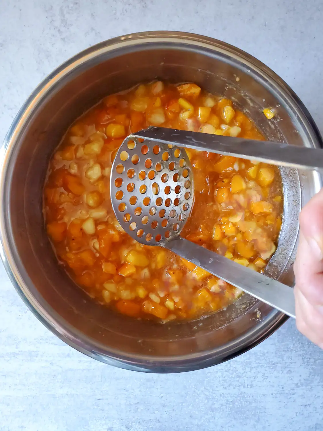 A stainless steel masher is resting over a stainless steel bowl full of macerated peaches that will turn into low sugar peach jam. Chunks of the fruit are still visible as they have not been fully broken down into jam consistency. 