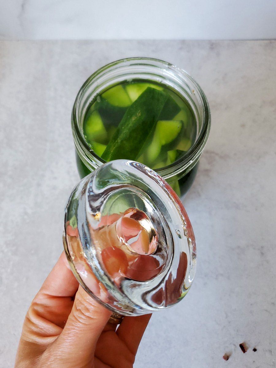 A hand is holding a glass ferment weight next to a mason jar of halved cucumbers. 
