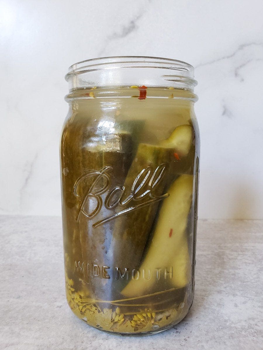 A quart mason jar full of halved cucumbers sitting in a ferment brine. Grape leaves, dill heads, and red chili flakes are inside the jar as well. 