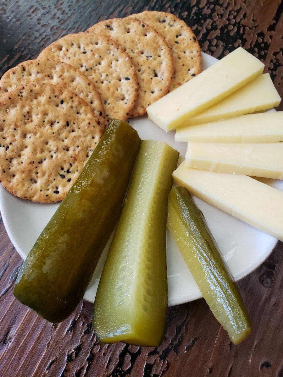 A small white plate is full of crackers, cheese, and fermented pickle spears, each taking up an equal amount of space on the plate. 