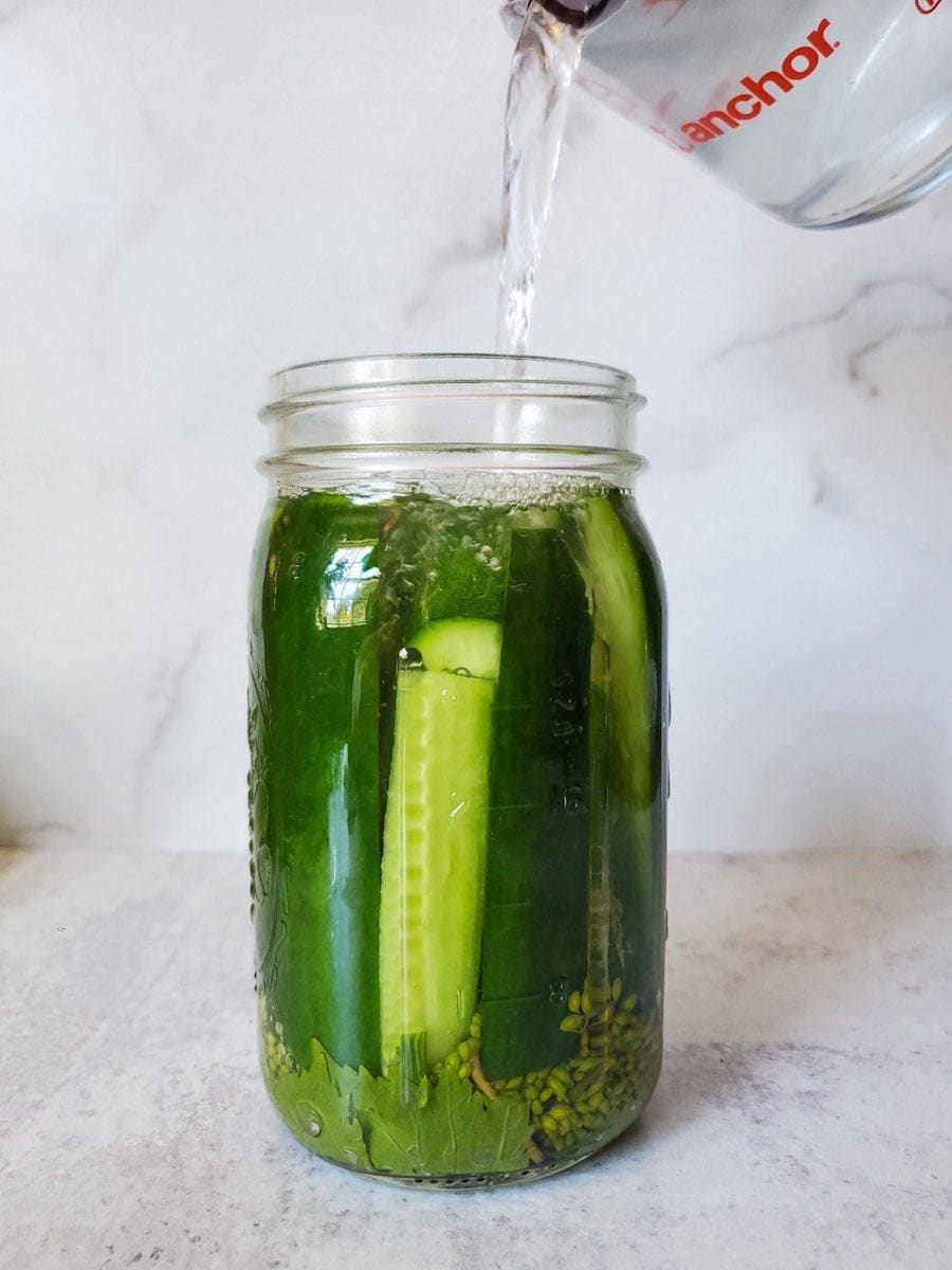 A quart mason jar is full of halved cucumbers that have been placed inside the jar vertically to pack it full. Grapes leaves and dill heads are visible on the bottom of the jar while a liquid measuring cup pours in the ferment brine from above to make the fermented pickles recipe. 