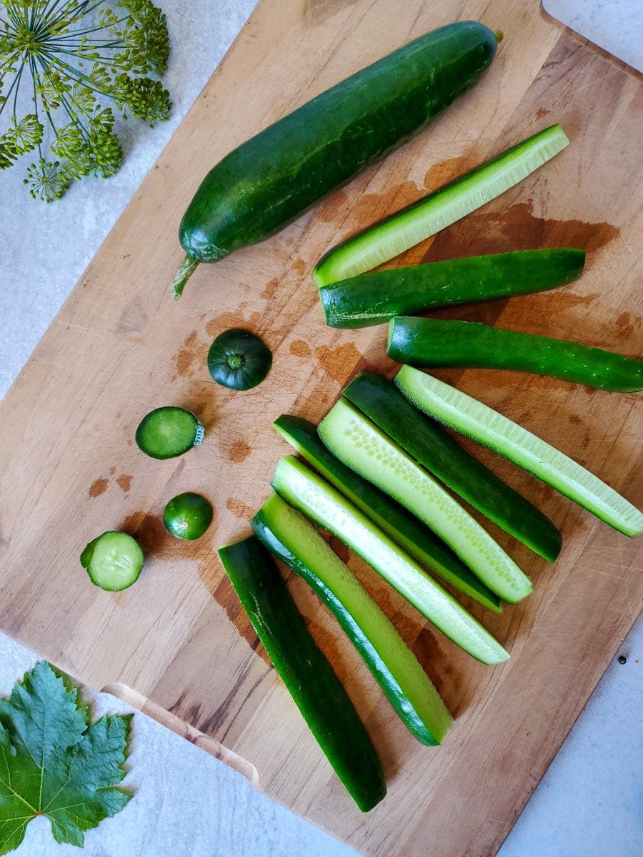 A flat lay of a wood cutting board with a whole cucumber on one side and cucumber quarters or spears that have been processed from whole cucumbers. A few of the discarded ends of the cucumbers are still on the board. These spears will be used in a fermented pickle recipe. 