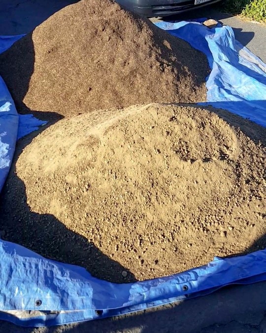 An image of two very large mounds of soil on a driveway, on top of a blue tarp. It was a delivery of several yards of bulk soil and organic compost.