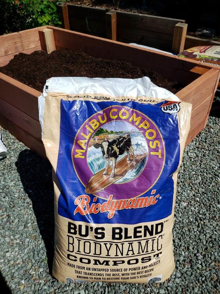 A photo of a bag of compost, by Malibu Compost. It is sitting on the gravel, leaning up against a half-filled raised bed behind it. 