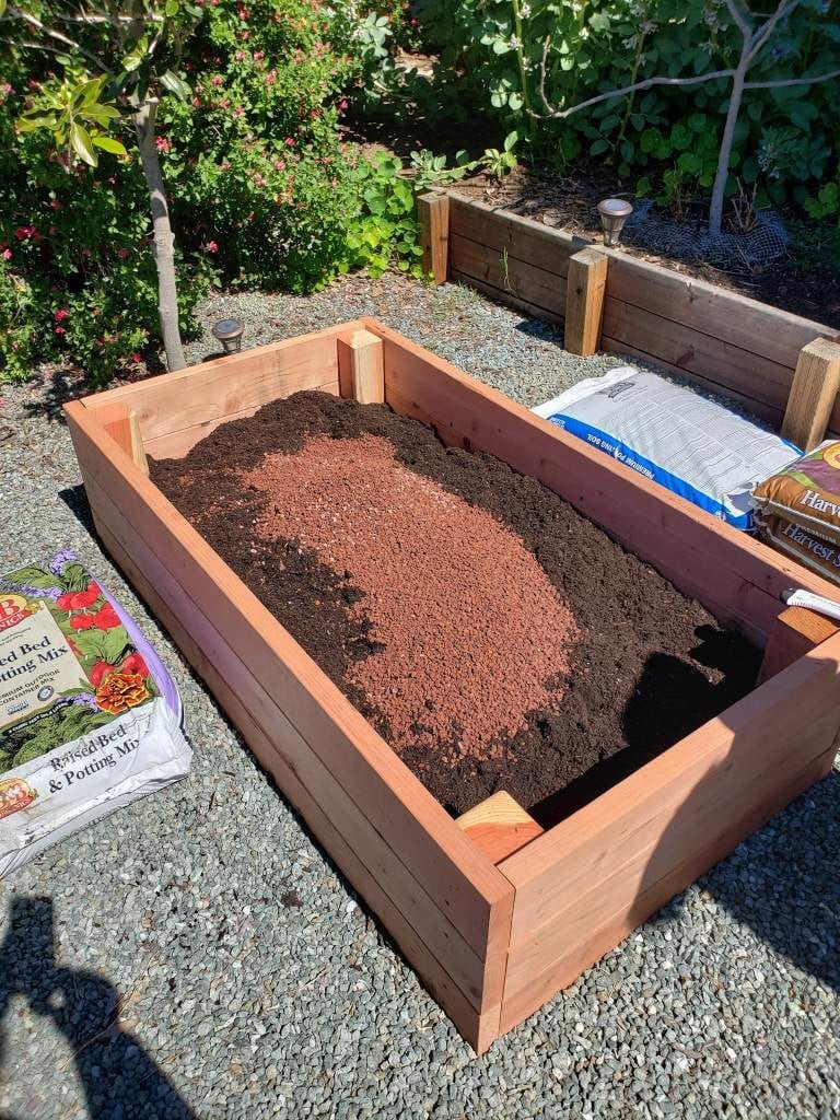 An image of a newly built redwood raised bed, only about half full at this time. In the bed, there is a combination of soil and compost, with some red volcanic rock on top, about to be mixed and then continued to be filled. 