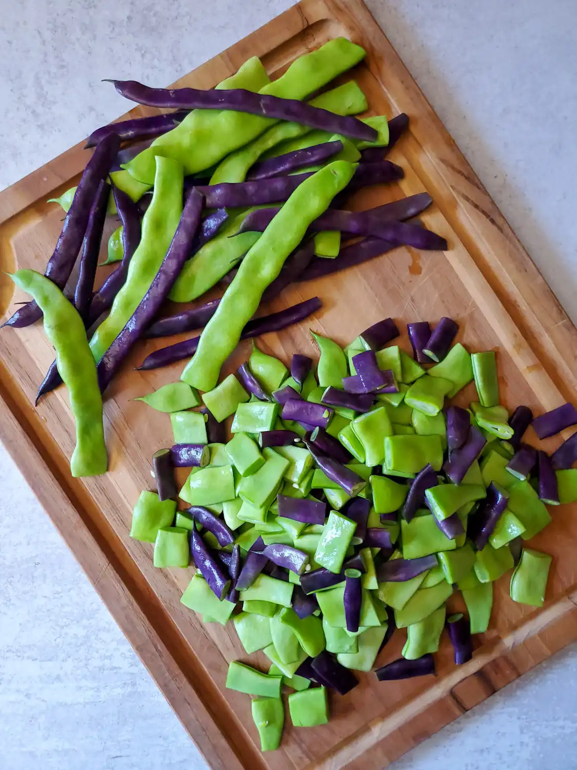 A wooden cutting board covered in purple and green veggies, half of them have been left whole while the other half have been cut into pieces. 