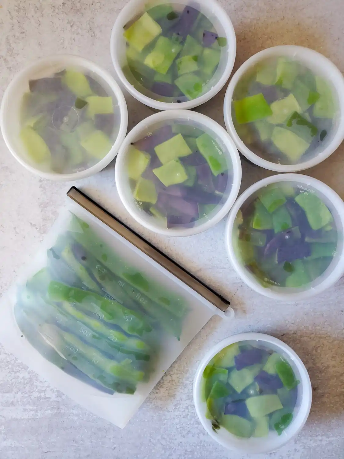 A flat lay image of 6 pints of blanched and cut green beans and one reusable silicone bag full of blanched and whole green beans. A variety of green and purple are showing through the containers from the colorful beans. 