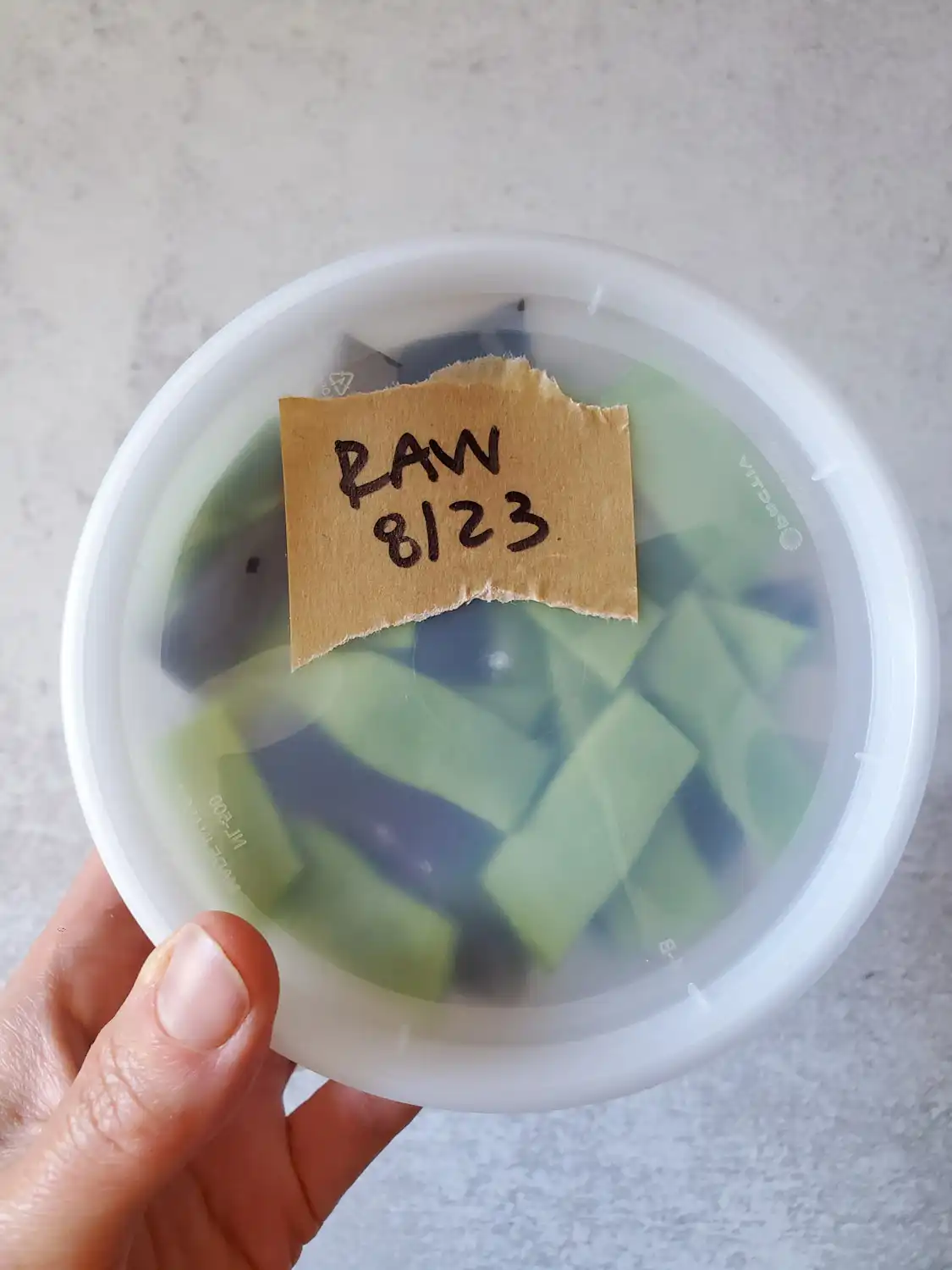 A flat lay of a freezer safe container containing raw cut green beans. There is a piece of brown tape on the lid with "Raw, 8/23" written on it in black marker. 