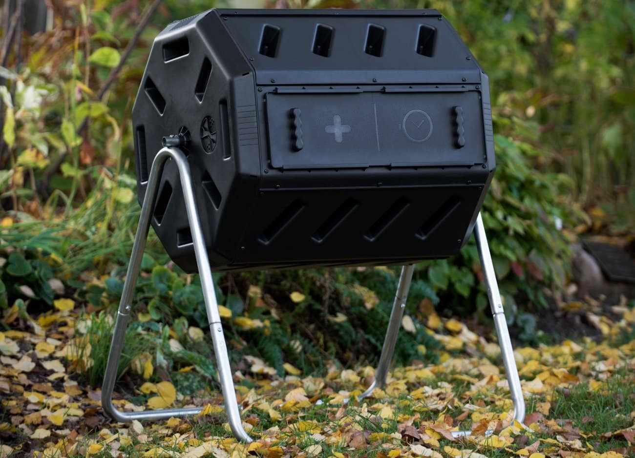 A black plastic compost tumbler sits amongst a wooded area. The tumbler contains two compartments so you can feed on side while letting the other side break down further. 
