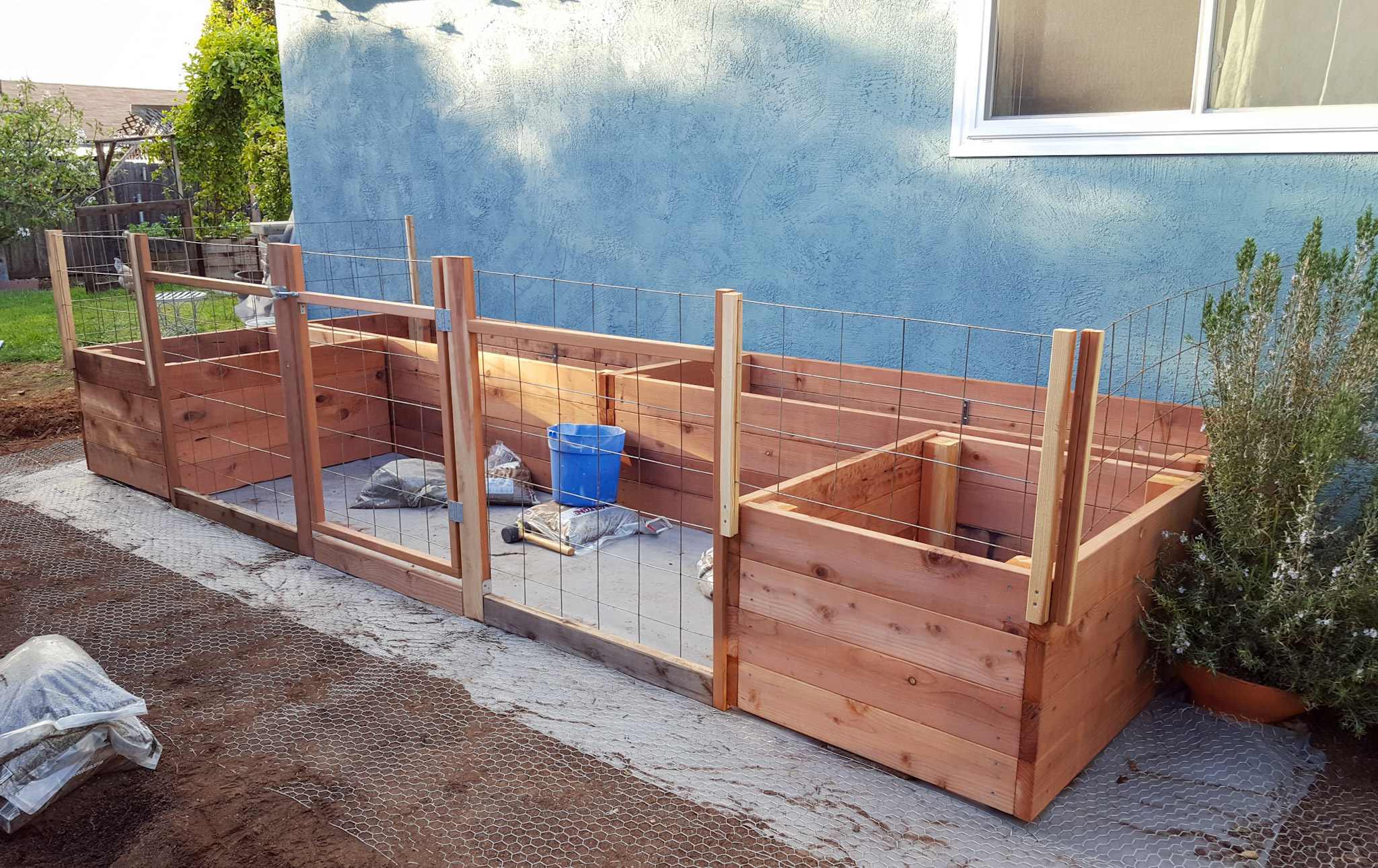 Four garden beds lined up against a house set in a U-shape. The beds are sitting on top of a layer of weed block fabric. There is fencing and a gate built into the front side of the garden beds where it would be open to chickens or other critters that is made with redwood 2x2's, 2x4's, and concrete remesh material. 