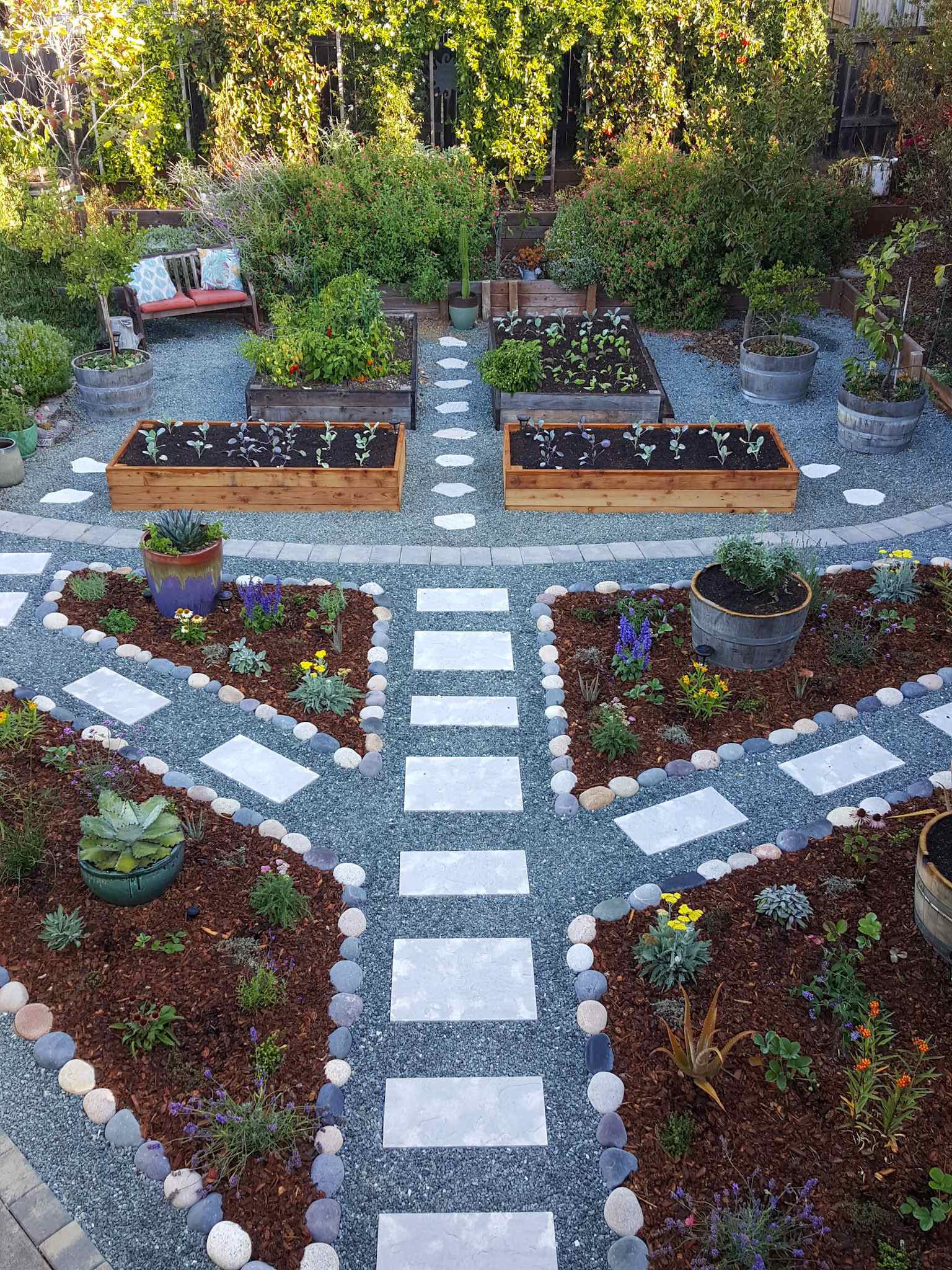 An image of the front yard taken from the roof of the house. All types of garden mulch are on display here from pollinator islands and garden perimeters mulched with redwood bark, paver lined pathways of rock gravel, and garden beds mulched with woody compost. There are plants dotting every area from flowering perennials, to fruit trees, shrubs, vines, vegetable plants, cacti, and aloe. 