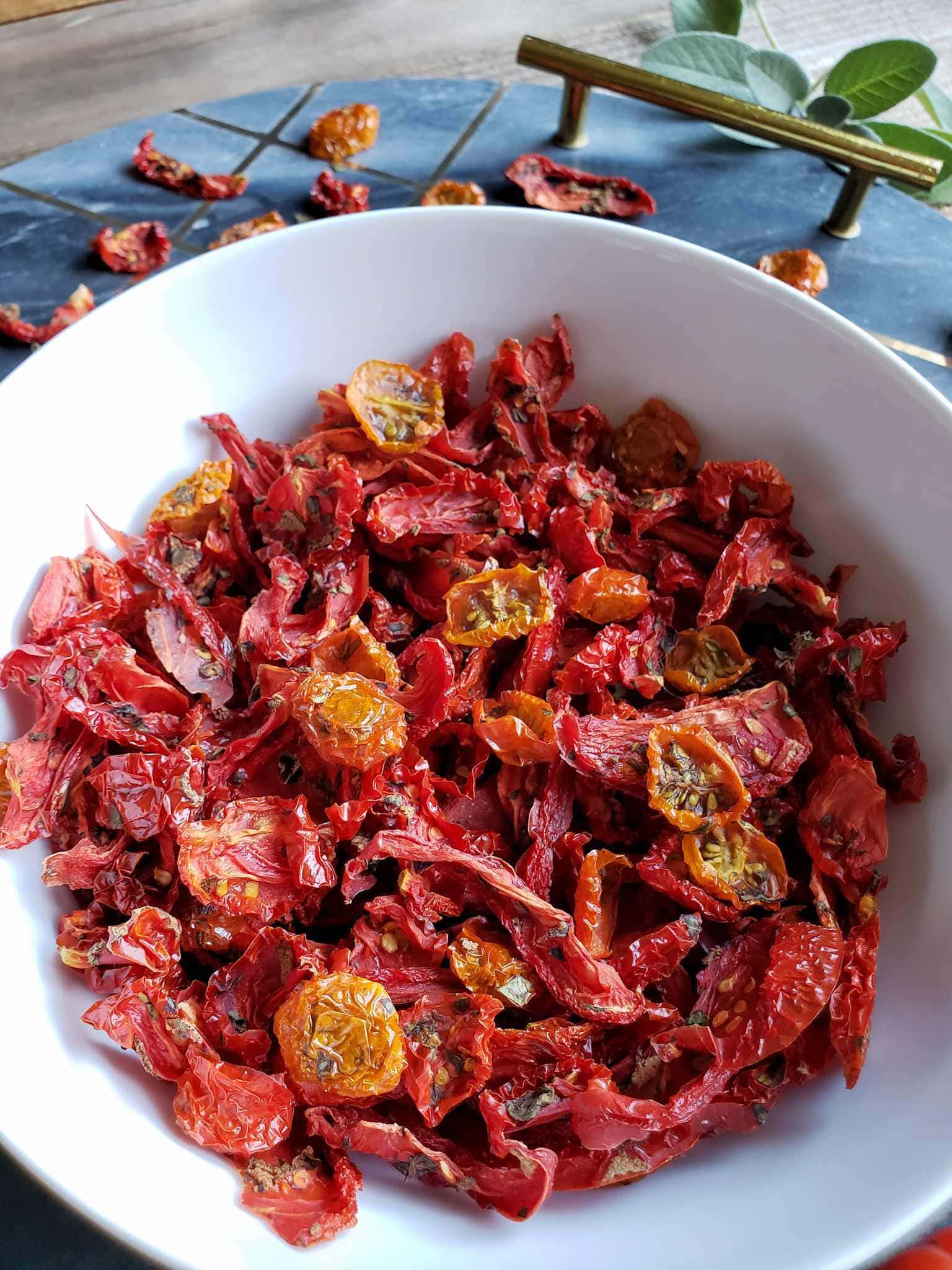 A close up image of a white ceramic bowl full of herb sun dried tomatoes, it is sitting on a round granite tray with handles, they are a few pieced of sun dried tomatoes scattered about and a luscious sprig of green sage.  