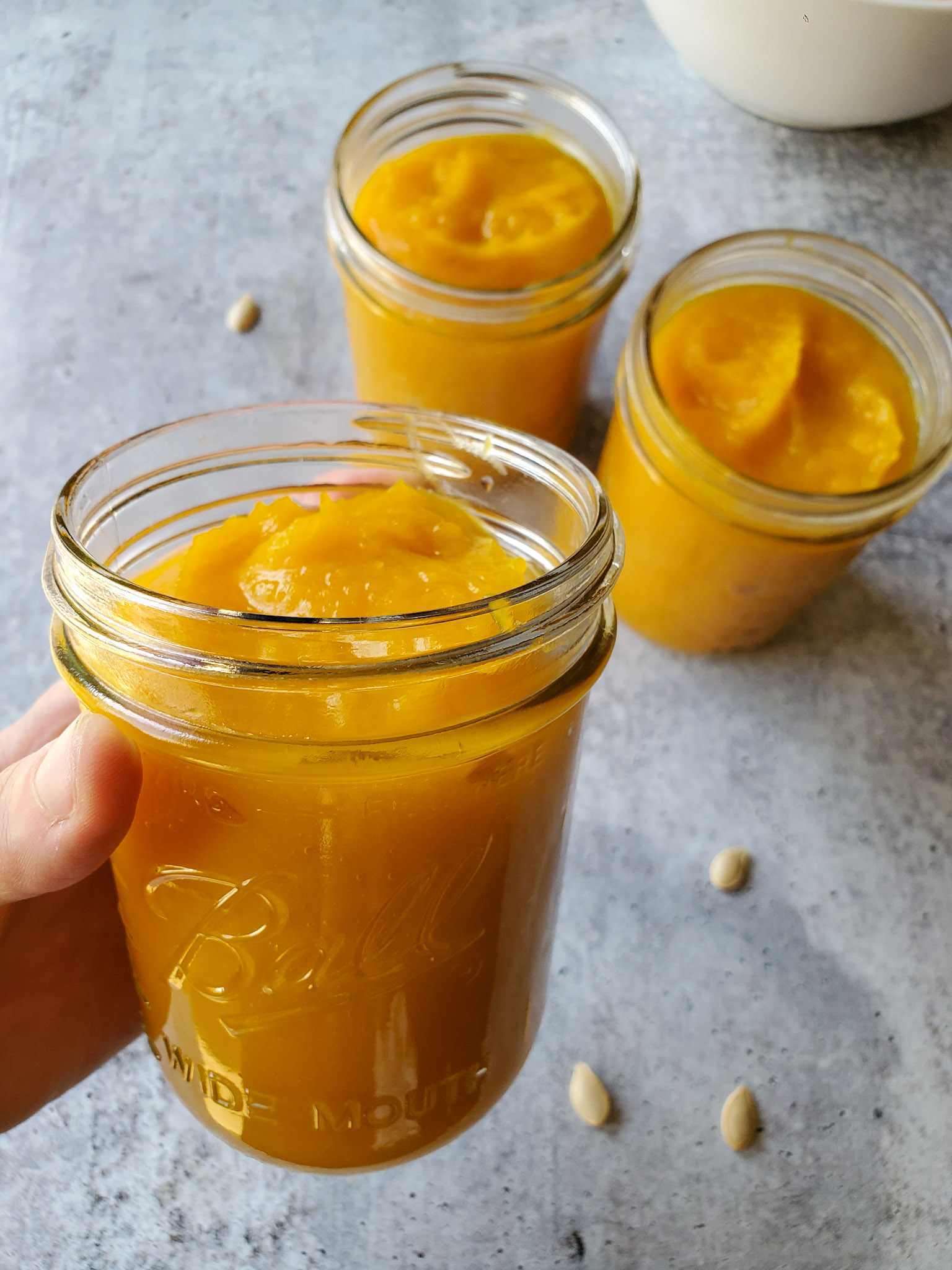 DeannaCat is holding a pint mason jar full of homemade pumpkin puree. It is vibrant orangish yellow in color, in the background there are two more pints of the pumpkin puree along with a few pumpkin seeds scattered about. 