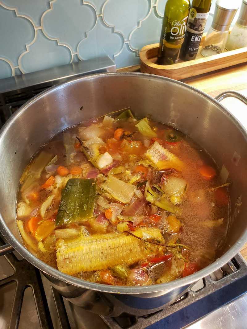 A stock pot full of vegetable broth scraps. There are corn cobs, leeks, carrots, onions, herbs, and garlic amongst other additions, all floating in the rich looking broth. 