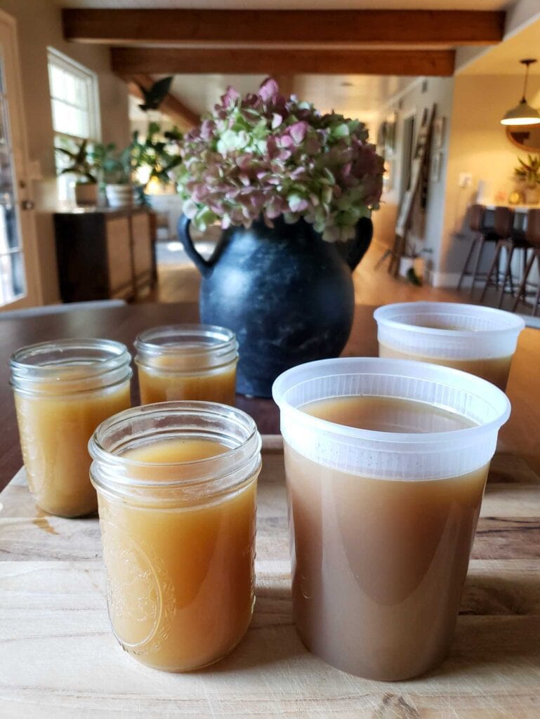A wooden cutting board sits atop a table, there are two BPA free plastic quart jars and three glass pint sized mason jars full of veggie broth.