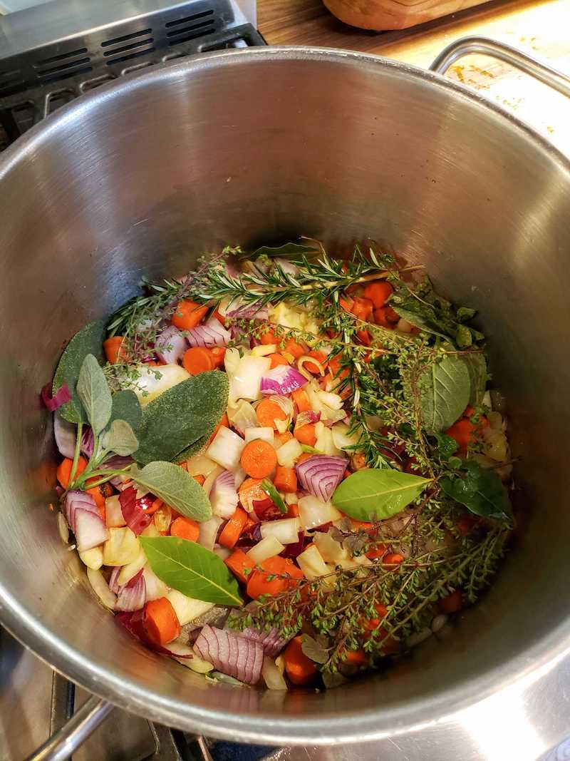 Fresh vegetable broth scraps are sitting in a stock pot ready to simmer on low for a minute before the frozen scraps are added. There are carrots, onions, an assortment of fresh herbs, and garlic visible. 