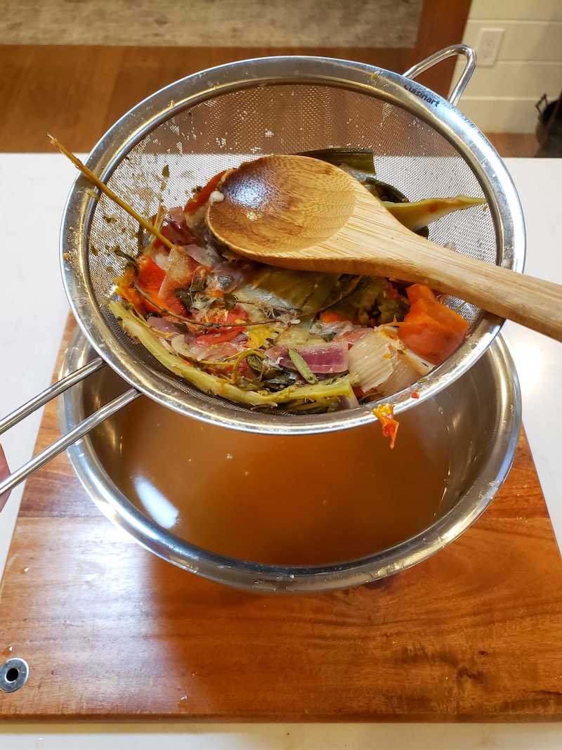 A metal strainer full of cooked vegetables sit over a metal bowl with brown liquid held within it. There is a wooden spoon resting on the scraps in the strainer as it was used to squeeze out the extra liquid from the veggies. 