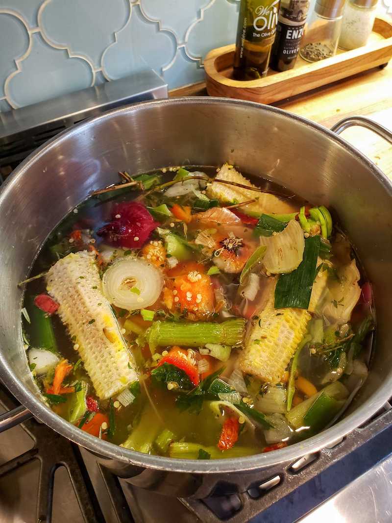 A stock pot full of vegetable scraps floating in water. Celery, corn cobs, bell pepper, onion, and carrots are visible.