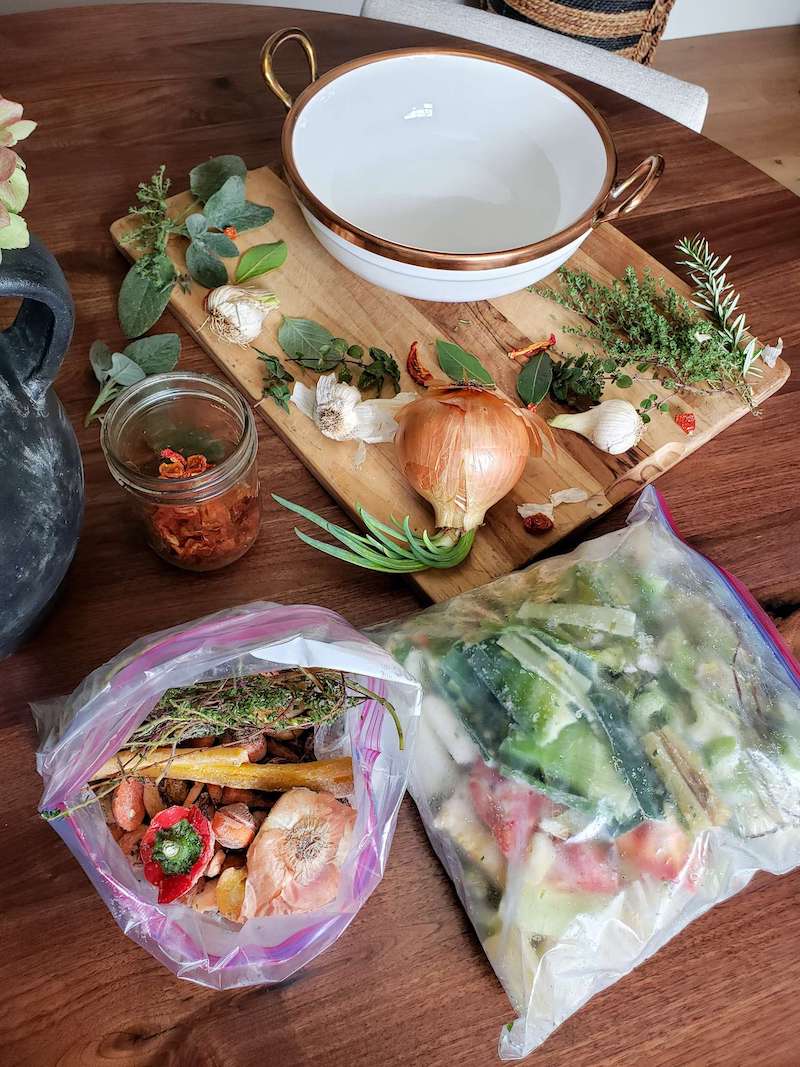 The ingredients for vegetable broth are laid out on a brown walnut table. There are two gallon sized freezer bags full of vegetable scraps of various kinds. On top of a cutting board there are fresh herbs, a sprouting onion, and a bulb of garlic next to a large white ceramic bowl. A mason jar is off to the side which is half full of sun dried tomatoes. 