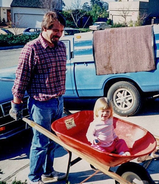 DeannaCat, a little blonde smiling girl, sitting in a red wheelbarrow at the age of two. Her dad is holding up the wheelbarrow, looking down at her, smiling. 