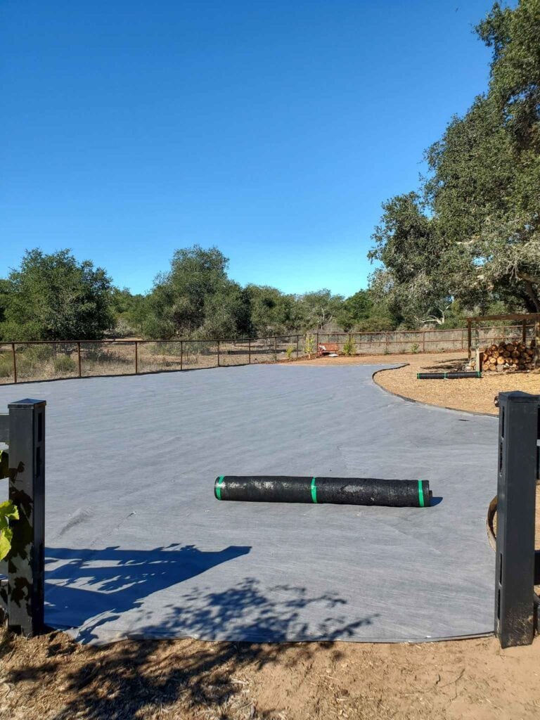 An open expanse of space has been covered with grey landscape fabric, a black roll of landscape fabric sits atop it which will be the second layer. Oak trees surround the garden area in the distance. 