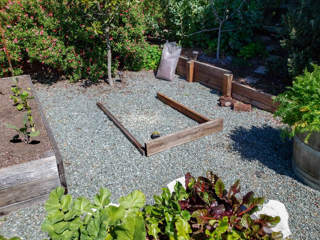 A few random boards lay in an open space in the gravel in a front yard garden, marking out where the future raised bed will go.