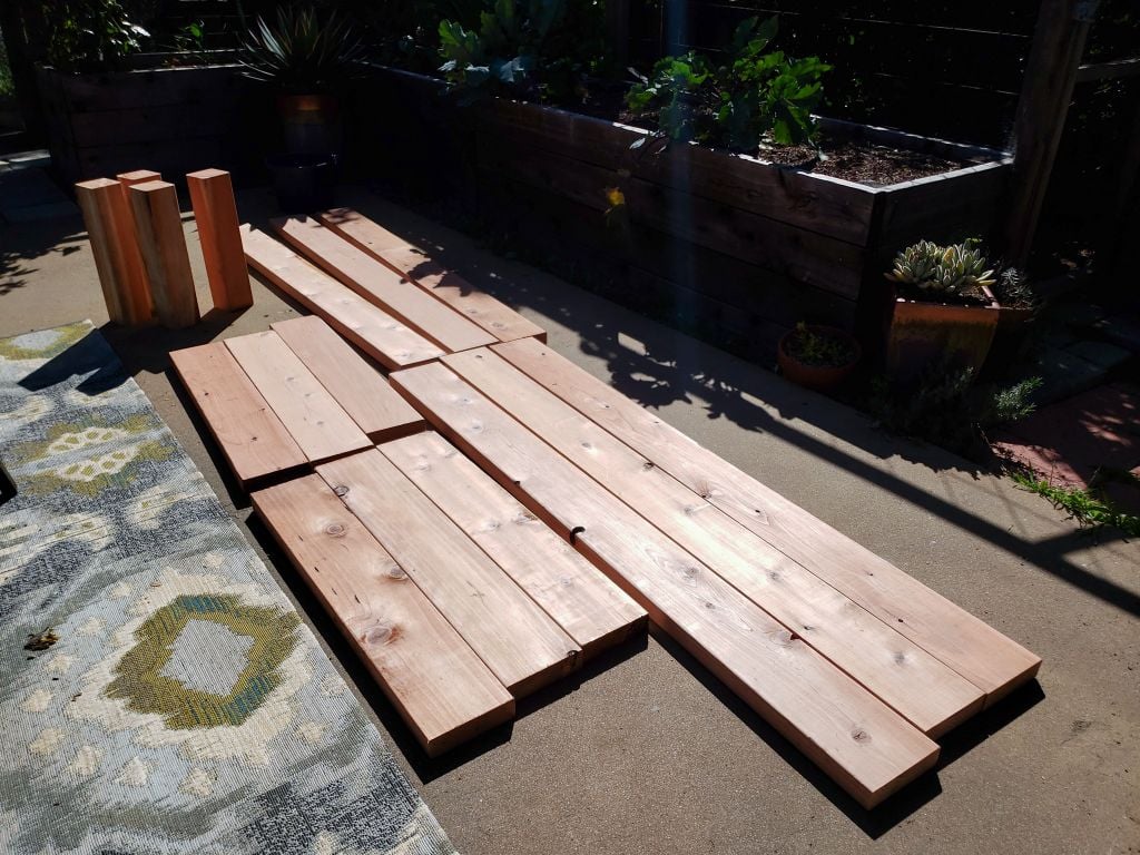 An image of a dozen cut redwood boards laying on a concrete patio, waiting to be put together into a raised bed. 