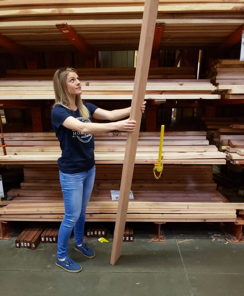 A photo of DeannaCat at home depot in the lumber section. She is a blonde woman with slender build, wearing blue jeans and a blue shirt, holding up a long redwood board vertically, peering up at it, standing in front of racks of boards.