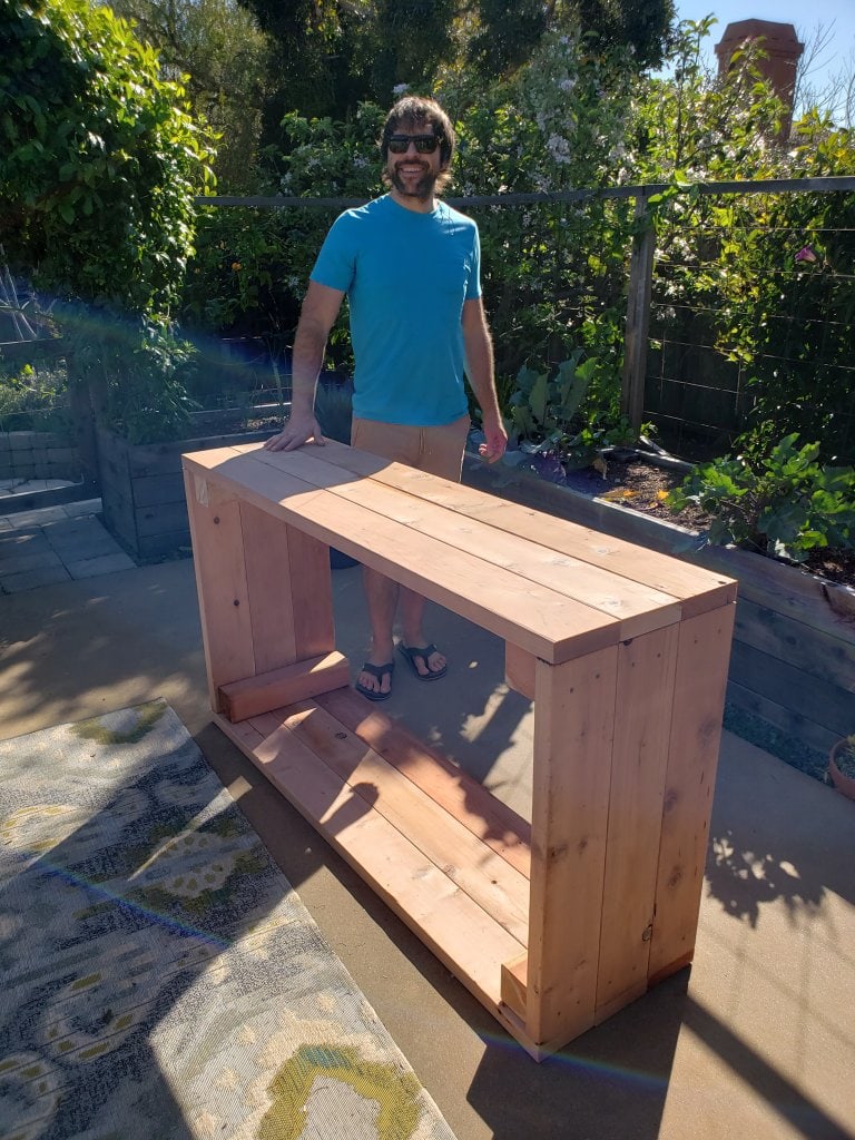 Aaron standing next to the just-built raised bed, still on the patio and on its side. He is smiling, wearing sunglasses and bright blue shirt, has dark brown hair, a short beard, and medium build. The patio garden and other raised beds (in use) are in teh background.