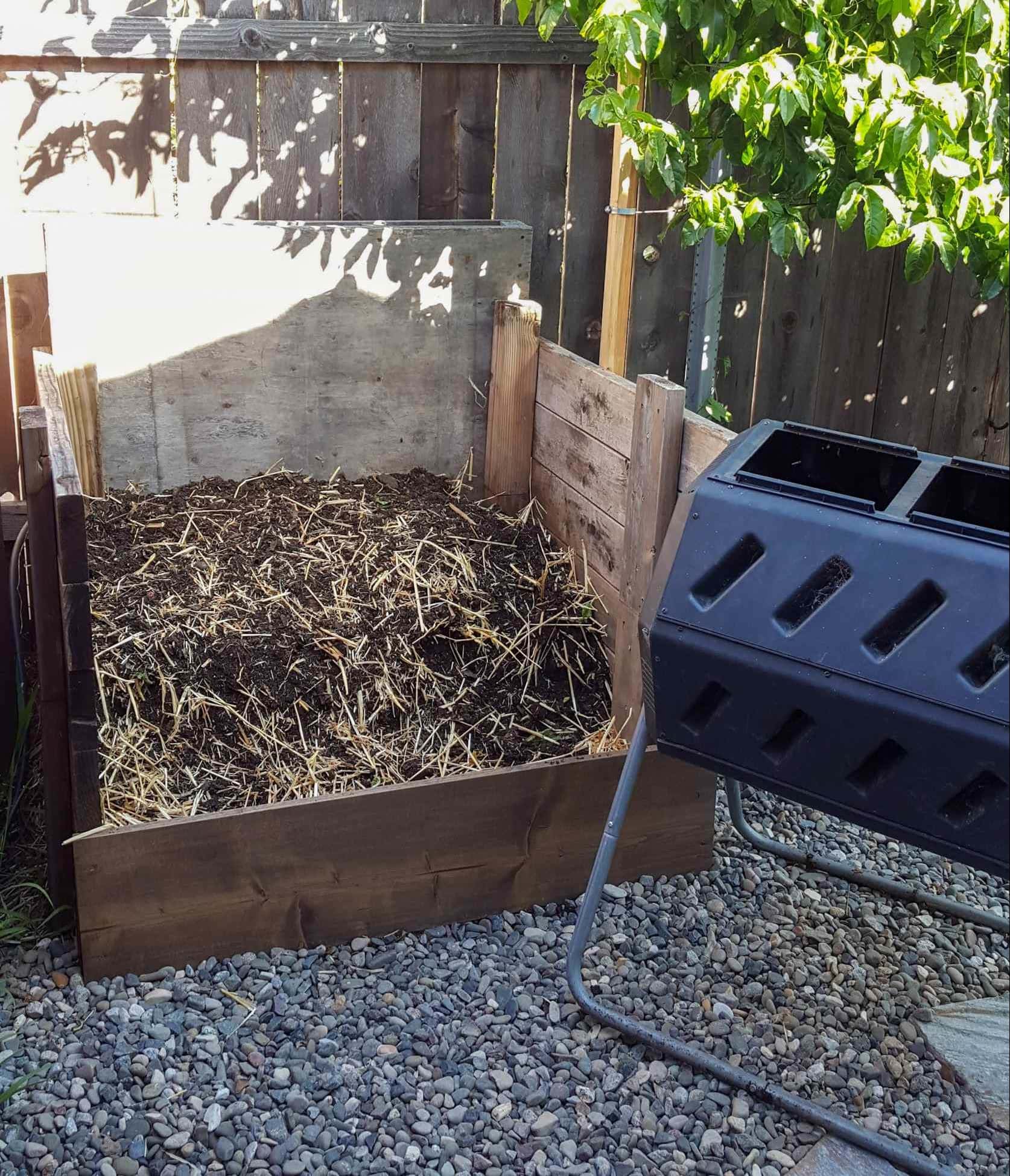 A three sided wooden compost stall is shown with a plastic compost tumbler staged next to it. The compost stall has some broken down material in it as well as some dried straw. These are  just two of the many options one can use to make compost.