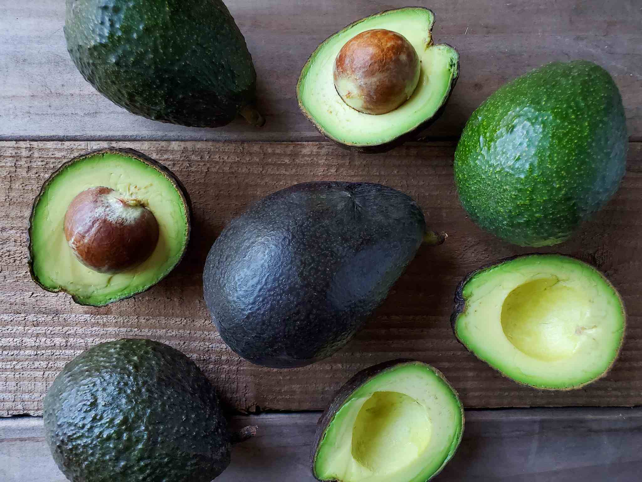 A number of whole and halved avocados are arranged on a wooden board. They each are at a varying stage of ripeness, some green, some dark green, while a large ripe avocado that is black in color sits in the middle. The avocados that have been cut in half reveal their creamy green fruit within.