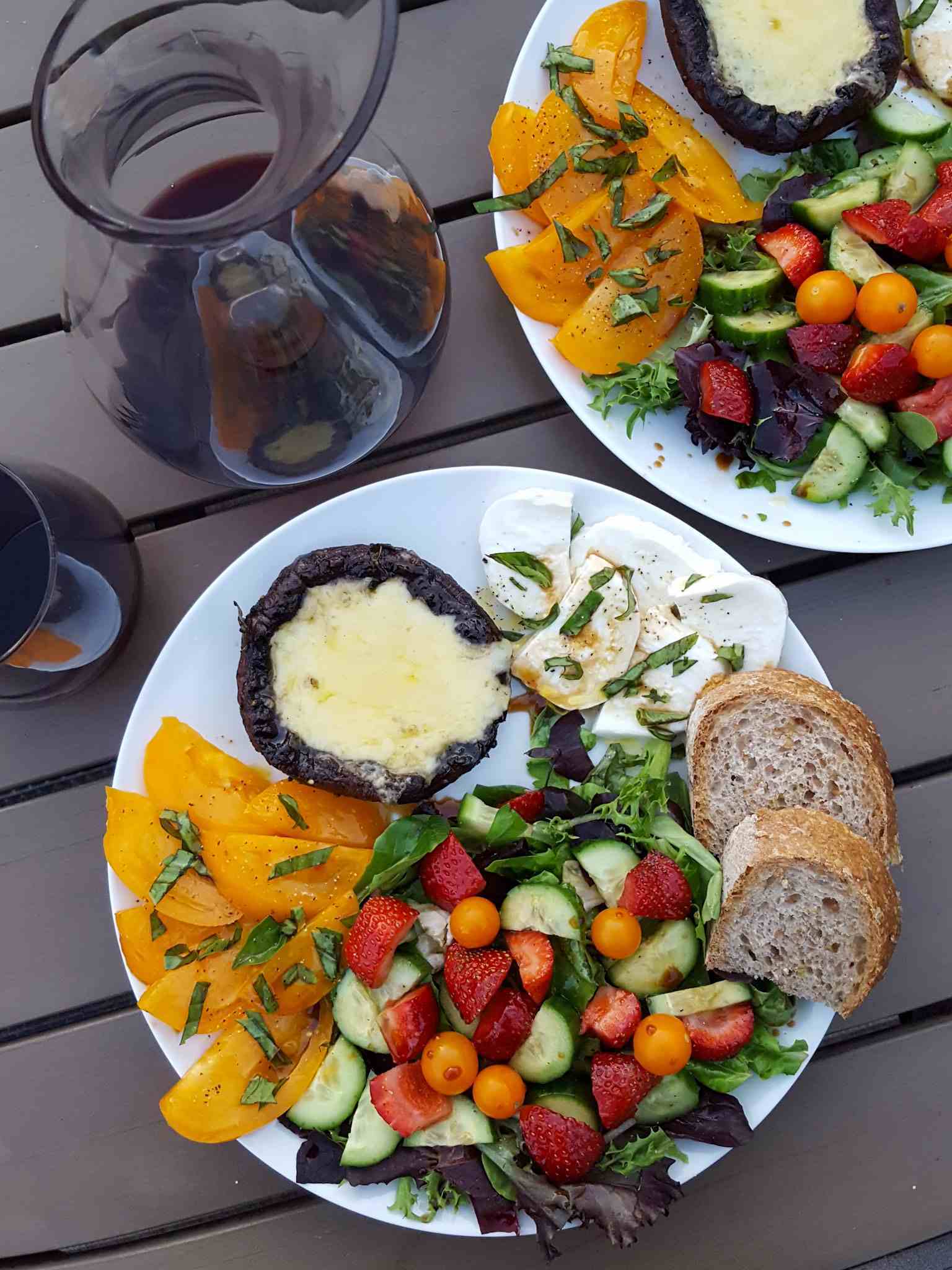 A mid summer dinner served on the outdoor patio table. There are two white ceramic dinner plates each one filled with fresh sliced yellow tomatoes, a green salad with Sungold tomatoes, cucumber, and strawberries, two slices of sourdough bread, slices of fresh mozzarella, and a grilled portabella mushroom with melted cheese. There is a carafe of red wine next to a stemless wine glass full of wine. 