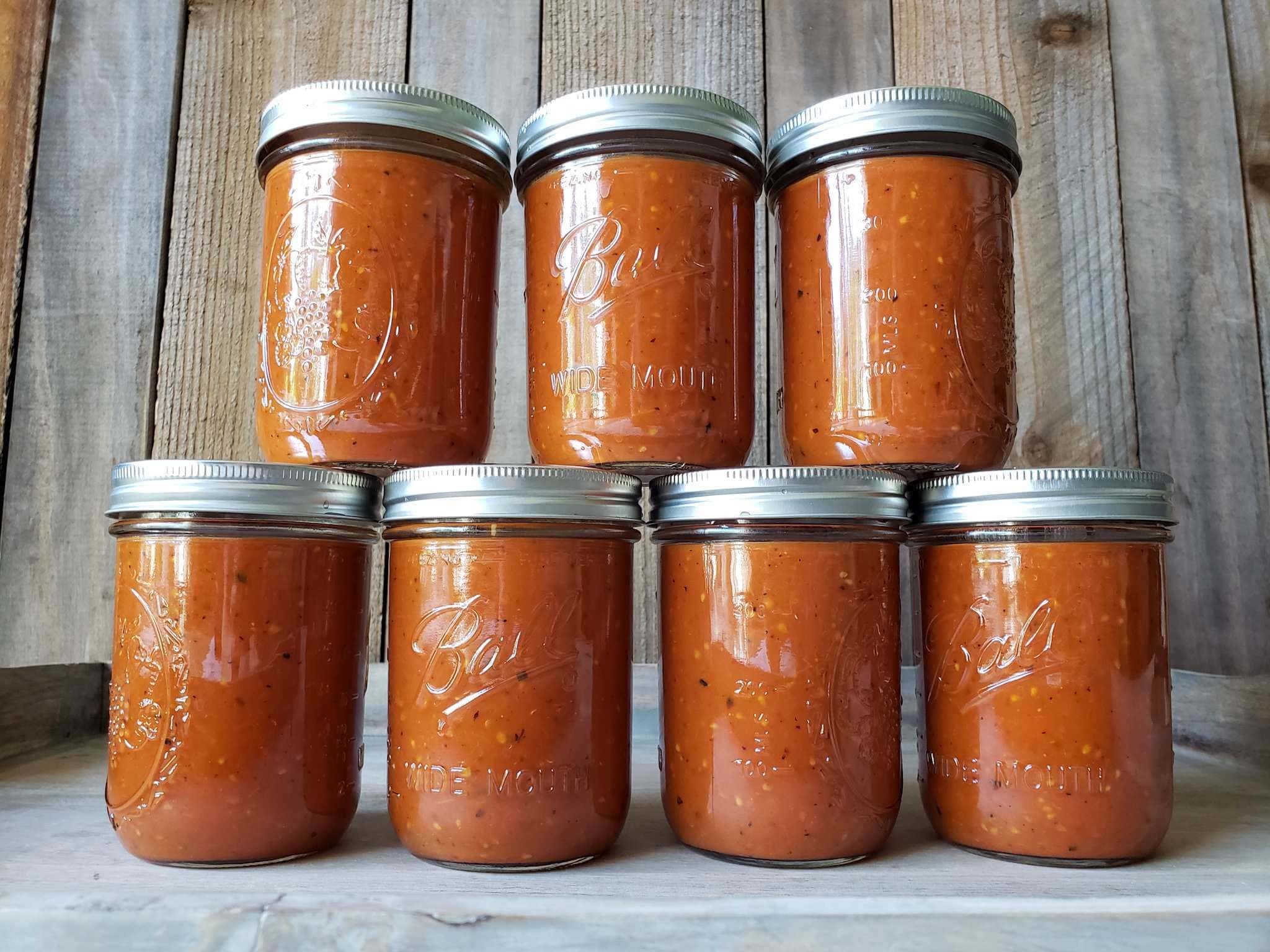 Seven pint jars of roasted tomato sauce are shown, four jars along the bottom with three jars stacked on top of the bottom four. The sauce is dark red in color with specks of black from the roasting. When you grow tomatoes, preserving them is a must to make the most of your harvest. 