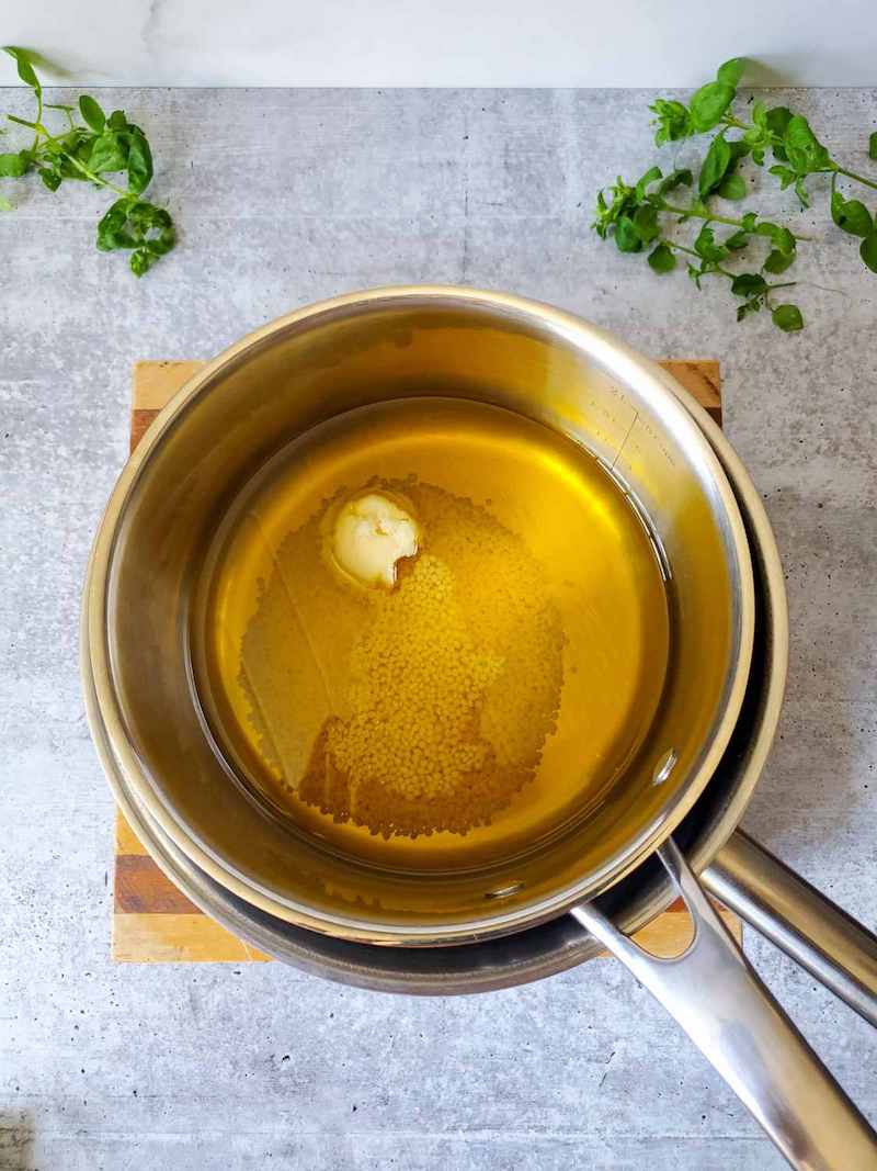 A stainless steel pot is sitting inside of a large pot to create a double boiler. There is oil, beeswax pastilles, and a glob of shea butter in the top pot. 