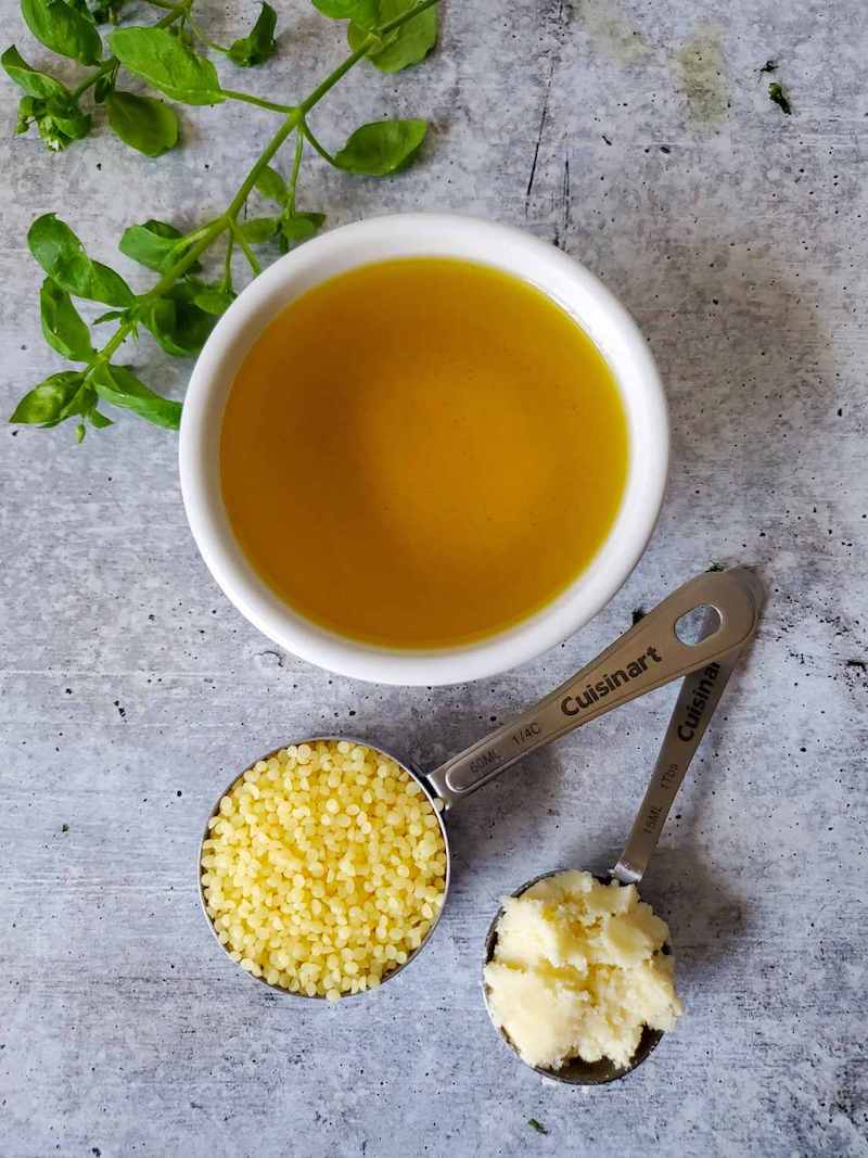 A white ramekin is full of oil sitting next to a metal measuring cup full of beeswax pastilles and a tablespoon measurement of shea butter. These are the ingredients to make chickweed salve. A fresh sprig of chickweed is splayed out as a garnish.